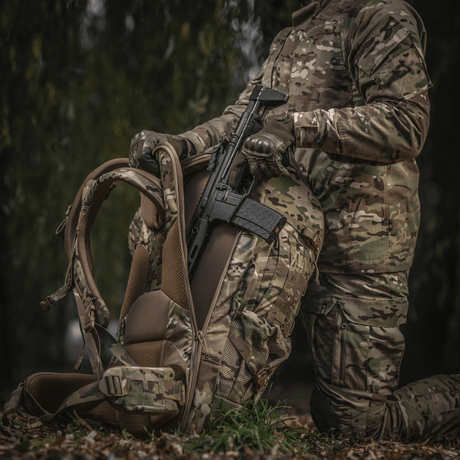 Henkilö pukeutuneena maastokuvioon pitää asetta ja M-Tac Large Gen.IV Elite -reppua metsäalueella.