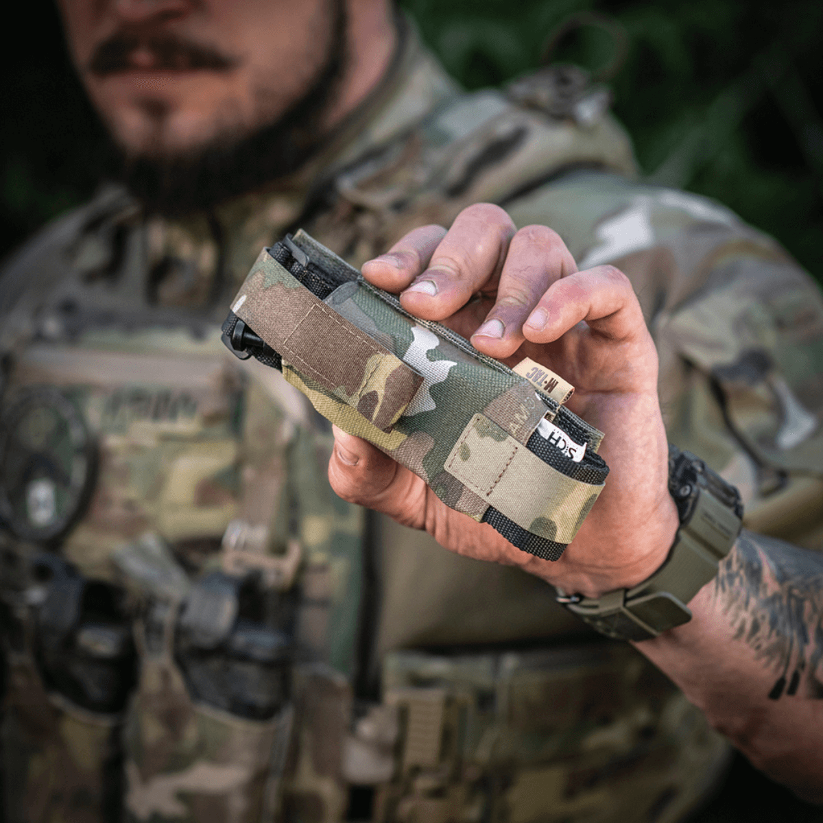 M-Tac Tourniquet Pouch Elastisk med MOLLE Gen.II i camouflage, holdt af person i militæruniform, hurtig adgang lomme til tourniquet.