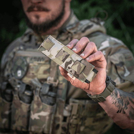M-Tac Tourniquet Pouch Elastisk med MOLLE Gen.II camo-designilla, pidetään taktisten varusteiden kantajan toimesta, näyttää helpon pääsyn ja säilytyksen.