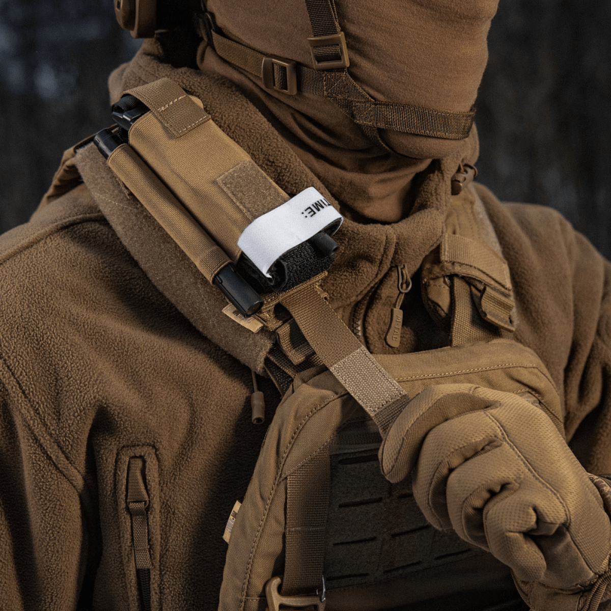 Person med brun beklædning bærer M-Tac Elastisk Tourniquet Pouch Molle Gen.III fastgjort til vest, udstyret med taktisk gear.