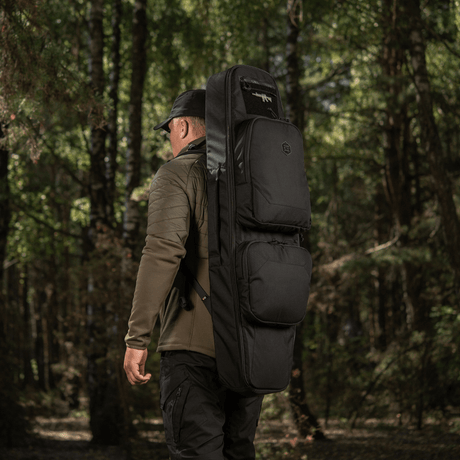 Mies kantaa M‑Tac Gun Backpack Case 105 cm Elite Hexiä metsässä, sopii AR-15- ja AK-kiväärien kuljetukseen. Cordura 500D -materiaali.