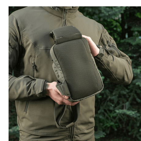 Henkilö esittelee M-Tac Sling Pistoltaske Elite Hexiä ulkona metsäympäristössä, takki vetoketjulla.