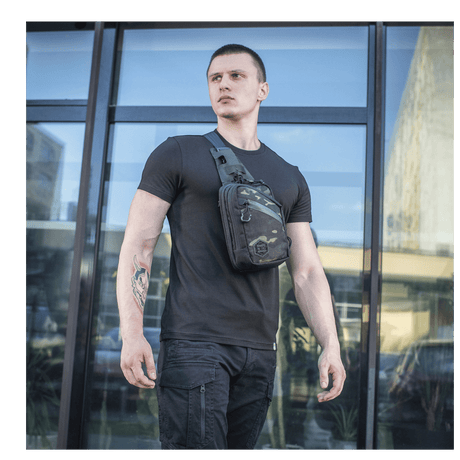 Mies M-Tac Sling Pistoltaske Elite Hex -laukulla lasirakennuksen edessä, näyttää monipuolisuutta ja tyyliä.