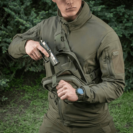 Henkilö, jolla on M-Tac Sling Pistoltaske Elite Hex, kantaa asetta metsätaustalla.