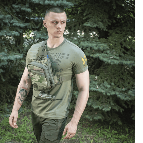Mies vihreällä M-Tac Sling Pistoltaske Elite Hex -laukkulla, valmistettu vahvasta Cordurasta, säädettävä hihna metsäalueella.