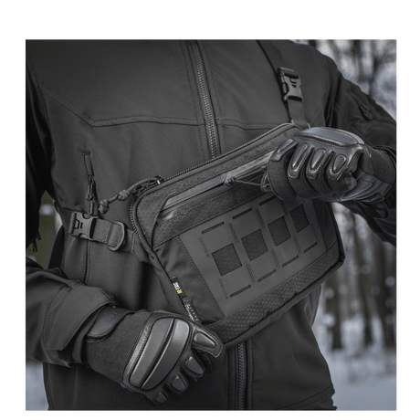 Henkilö mustassa takissa esittelee M-Tac Admin-laukku Eliteä, valmistettu kestävästä Cordura-materiaalista, vetoketjuilla ja korkealaatuisilla soljilla
