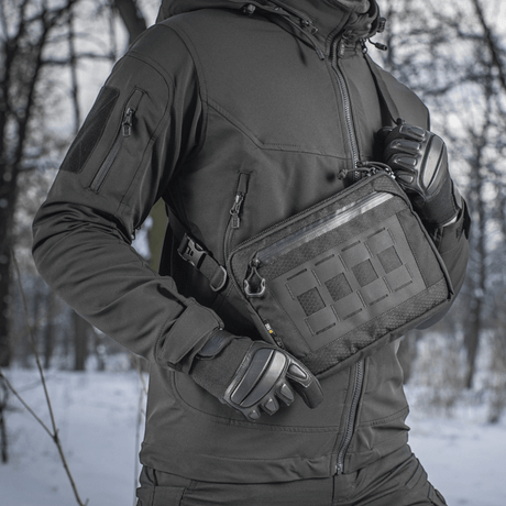 Henkilö pukeutuneena mustaan takkiin M-Tac Admin-laukku Eliten kanssa, valmistettu kestävästä Cordura-materiaalista, lumisessa taustassa