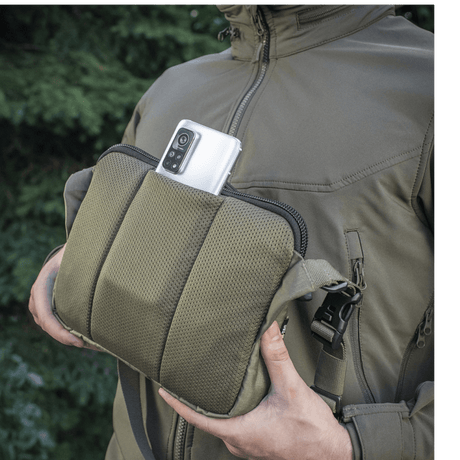 M-Tac Admin Taske Elite Cordura-kankaasta, älypuhelin taskussa, säädettävä olkahihna ja tukeva rakenne, ulkoilmataustalla.