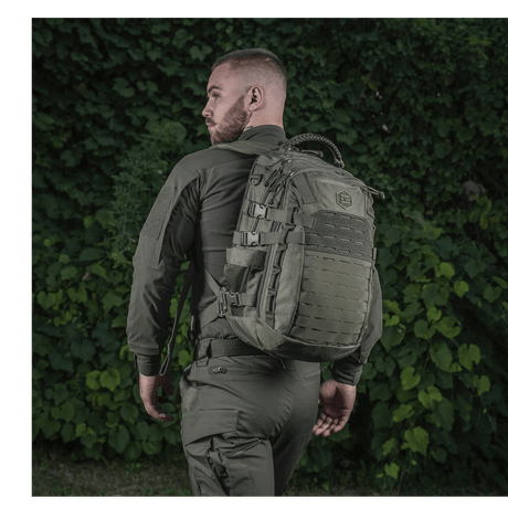 Henkilö M-Tac Mission Rygsæk Elite Hex -repun kanssa, taustalla vihreää kasvillisuutta.