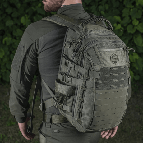 M-Tac Mission Rygsæk Elite Hex henkilön selässä taktisten vaatteiden kanssa, Cordura-materiaali ja MOLLE-järjestelmä, nähtynä takaa luonnossa.