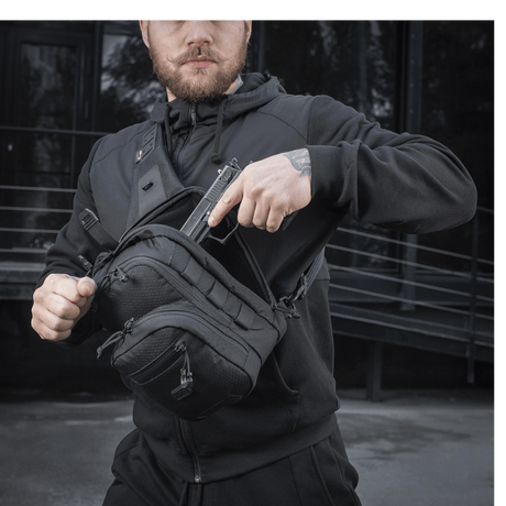 Mies käyttää M-Tac Cross Taske Slim Elite Hex -laukkua mustana Cordura-materiaaleilla, vetoketjuilla ja YKK- ja Woojin-soljilla, ulkoilmassa.