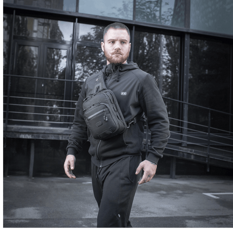 Mies M-Tac Cross Taske Slim Elite Hex -laukku olallaan, pukeutunut mustiin vaatteisiin modernin rakennuksen edessä.