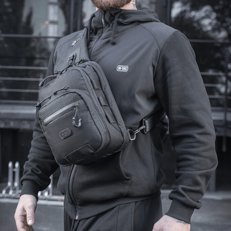 Henkilö kantaa M-Tac Cross Taske Slim Elite Hex -laukkua, pukeutunut mustiin, ulkona.