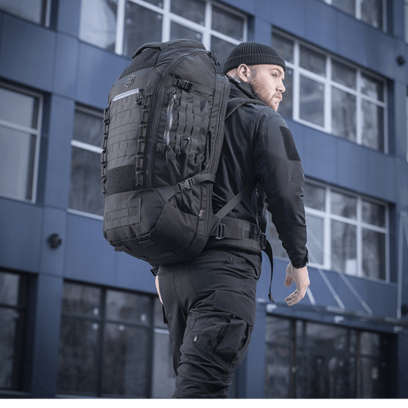 Mies kantaa M-Tac Rygsæk Large Elite Hex Gen. IV -reppua mustana, ergonomisesti suunniteltu toiminnallisuuteen ja kestävyyteen, nähty kaupunkiympäristössä.