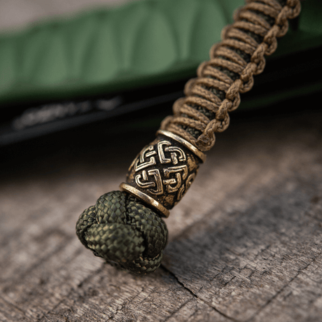 M-Tac Knife Lanyard Celtic, punottu paracord kelttikuvioinnilla, veitsilisävaruste puumaisella pinnalla.