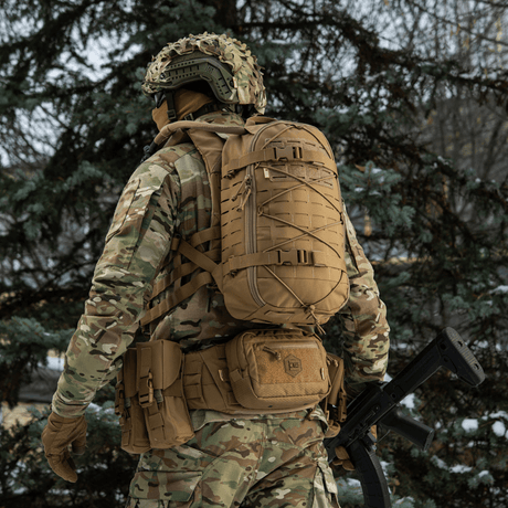 M-Tac reppu Sturm Elite Gen.II kentällä, mies maastopuvussa ja taktiikkareppu MOLLE-järjestelmällä ja vahvistetuilla remmeillä, metsäympäristö.