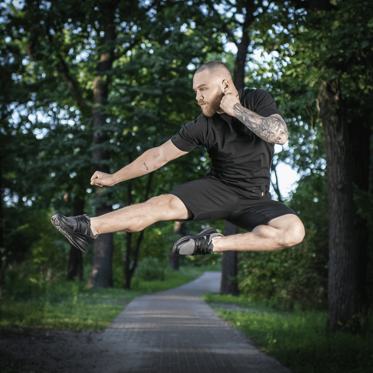 Mand i M-Tac Shorts Casual Fit Cotton udfører et spring i parken, iført afslappet sort tøj, fremstillet af åndbart 100% bomuld.