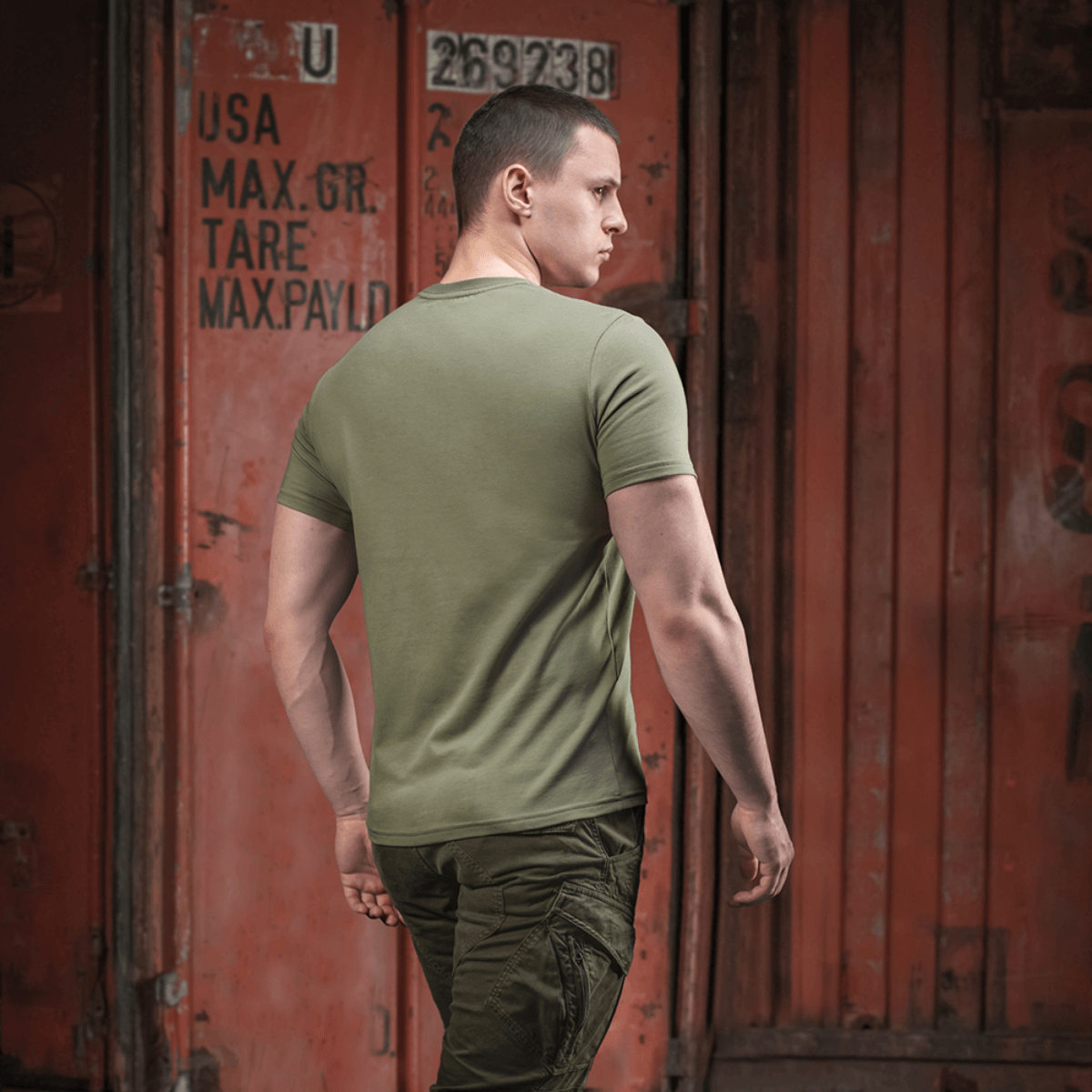 Person iført armygrøn M-Tac T-shirt Viking med ryggen til, gående forbi container i bymiljø.