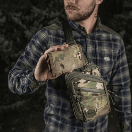 Mies pitelee M-Tac Pung Elite Gen.II:tä maastokuvioisena, valmistettu Cordura 500D:stä ja varustettu YKK-vetoketjuilla, metsäympäristössä.
