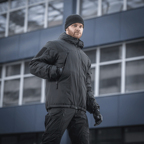 Mand iført M-Tac Alpha Gen.III Pro Vinterjakke i sort, velegnet til temperaturer ned til -20° C, foran moderne bygning.