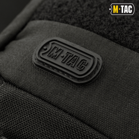 Suljettu lähikuva M-Tac-logosta M-Tac Buckler Taske Elite -laukun päällä, musta taktinen laukku MOLLE-järjestelmällä ja piilotetulla asekotelolla.