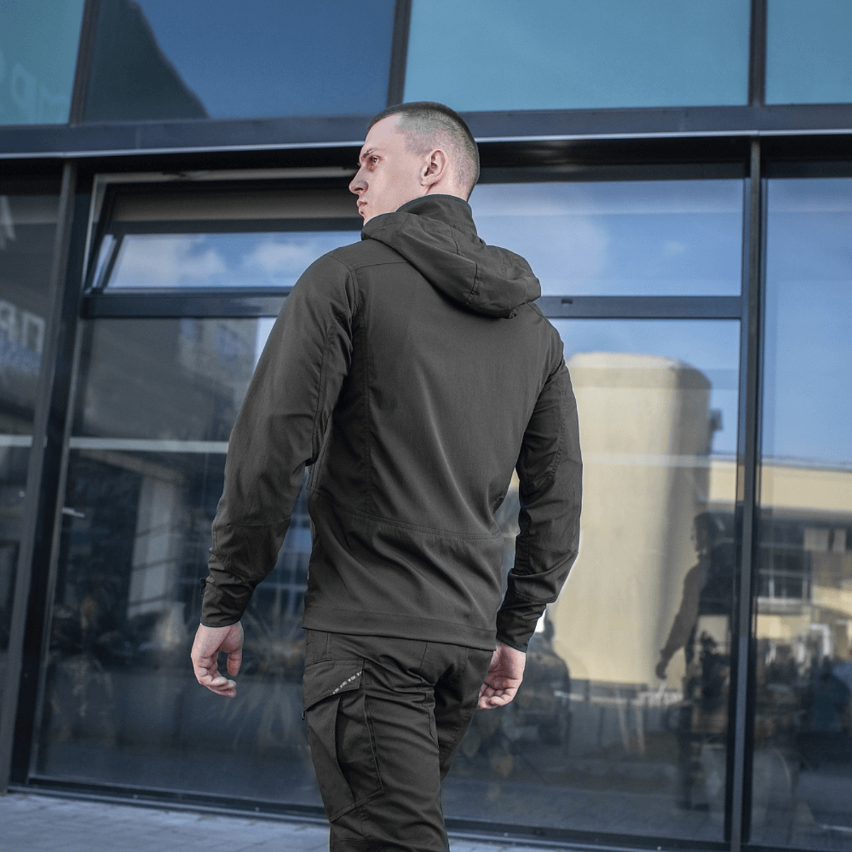 Mand iført sort M-Tac Flash Jakke, perfekt til vandreture og hverdagsbrug, set bagfra foran moderne bygning.