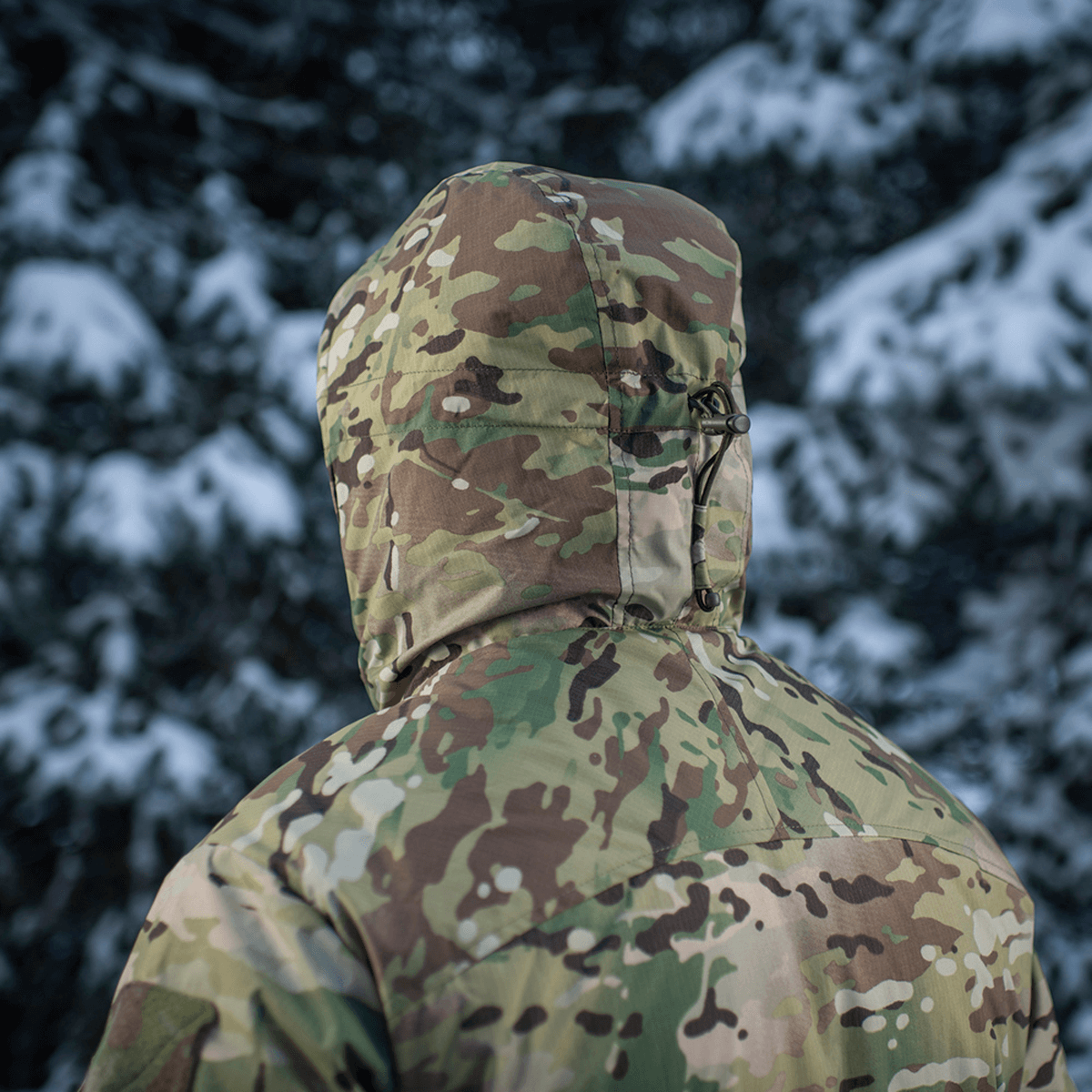 M-Tac Alpha Gen.IV Pro Primaloft Vinterjakke i Multicam stof mod snefyldte træer, ideel til kolde vejrforhold.