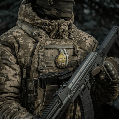 Sotilas maastokuvioisessa takissa kantaa M-Tac Patch M67 Kranaatti 3D PVC:tä liivissä, humoristinen emoji-kuvio, taktinen varustus.