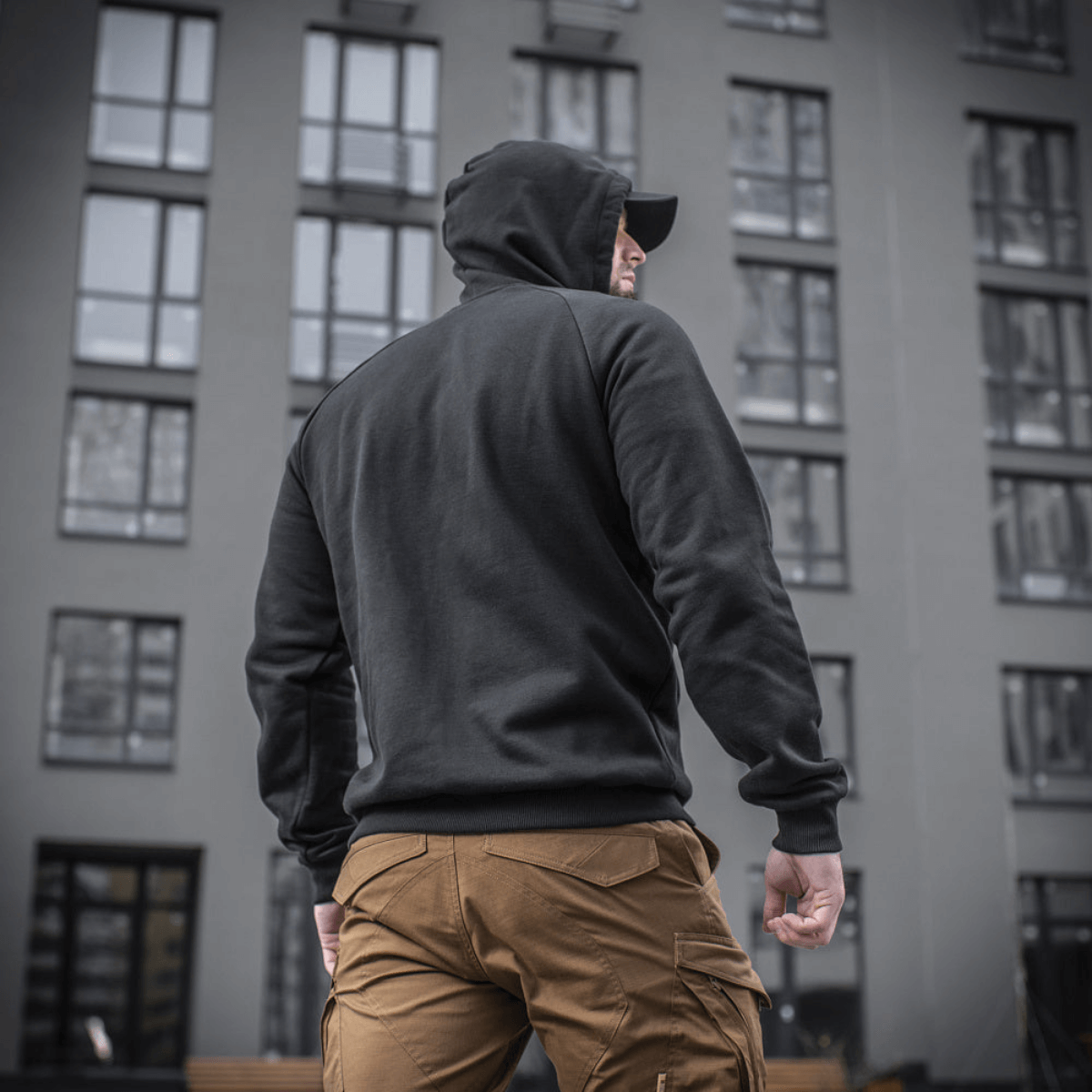 Mand i M-Tac Cotton Raglan Hard Hoodie, 100% bomuld, fleecefor, perfekt til efterår og vinter, set bagfra foran bygning.
