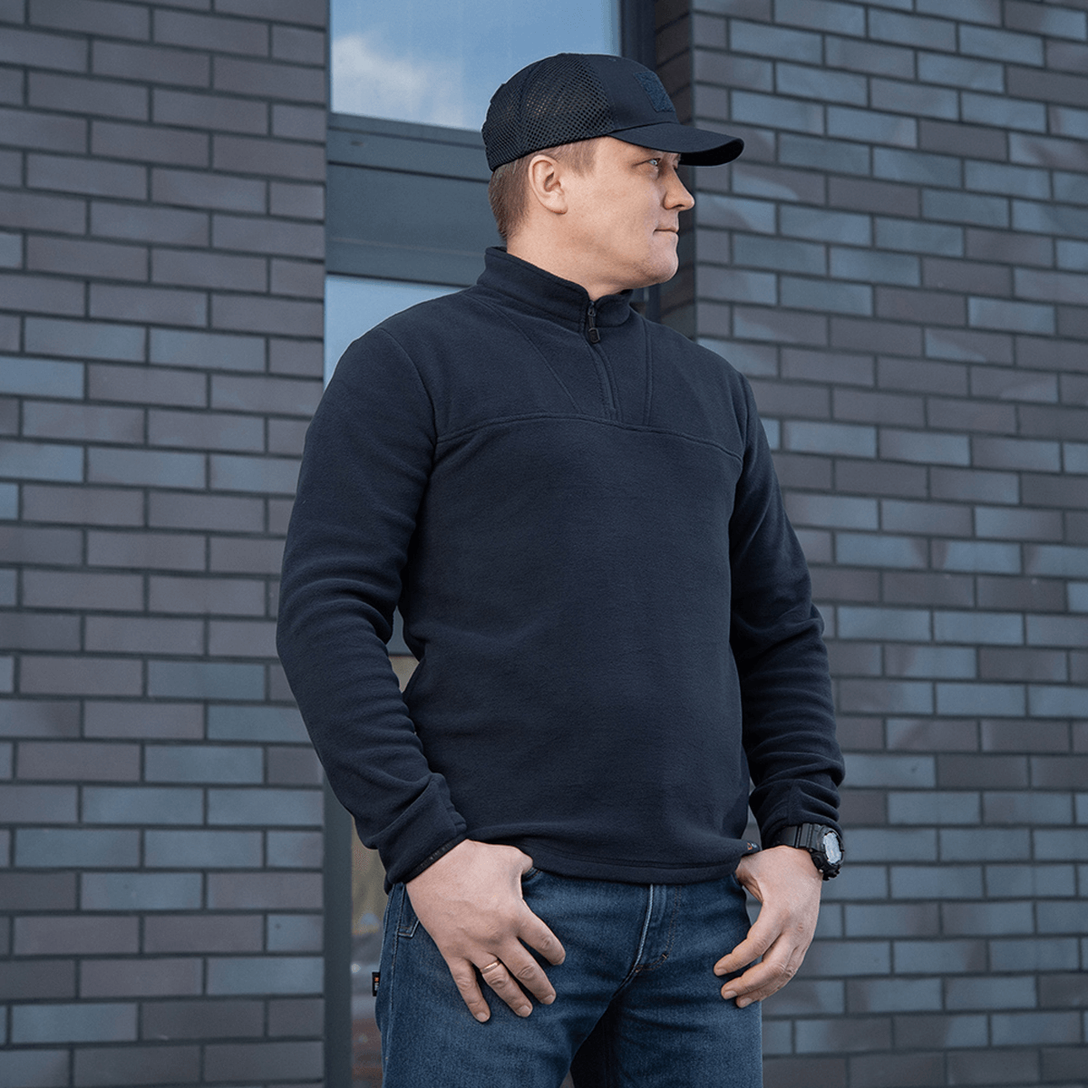 M-Tac Delta Polartec Fleecejakke i sort, fremstillet af Polartec®-fleece, bæres af en mand foran en murstensvæg.