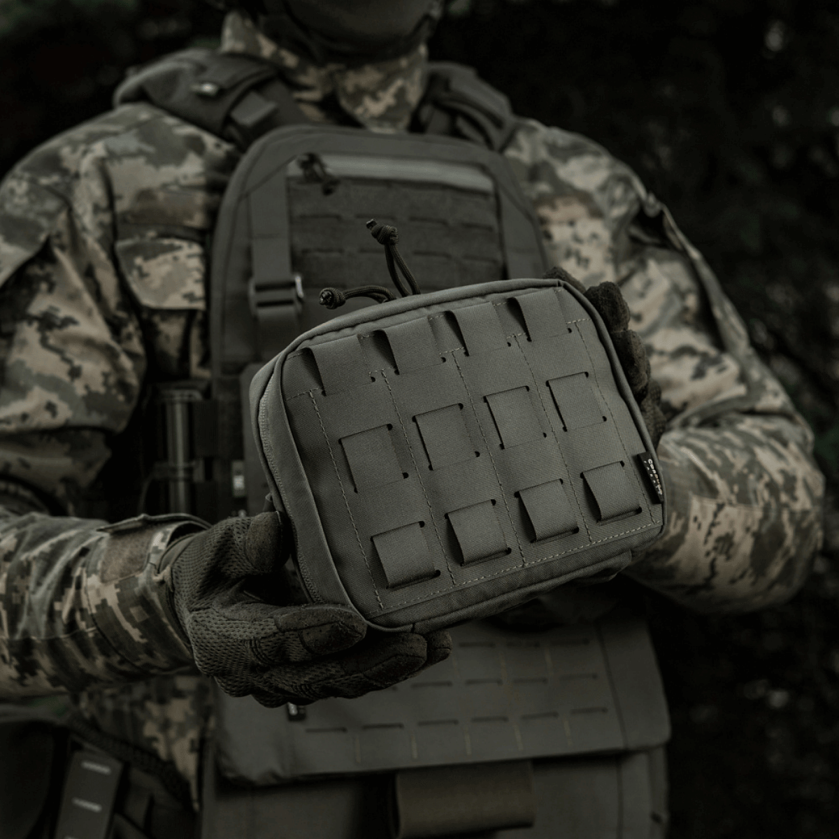 M-Tac organizer pouch Elite holdt af en taktisk klædt person, fastgjort til vest med M.O.L.L.E.-system, velegnet til førstehjælp og EDC-udstyr.