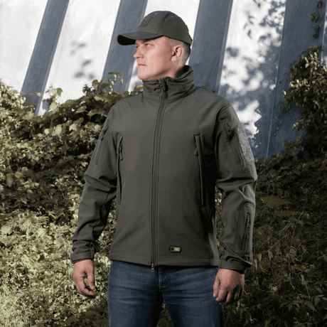 Mand iført M-Tac Soft Shell Jakke med fleecelag og vejrbeskyttelse, står udendørs foran grøn baggrund.