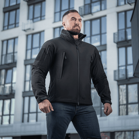 Mand iført M-Tac Soft Shell Jakke står foran en moderne bygning, viser jakkens vejrbeskyttende og isolerende egenskaber.