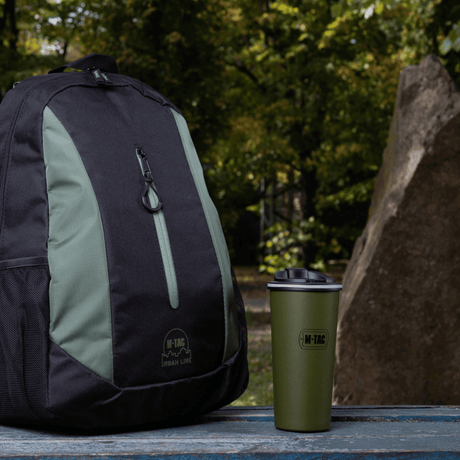 M-Tac 450 ml termosmuki venttiilillä pöydällä repun vieressä metsäkohtauksessa