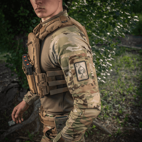 Mies maastokuvioisissa vaatteissa, M-Tac Patch Ace of Spades käsivarressa luonnonympäristössä.