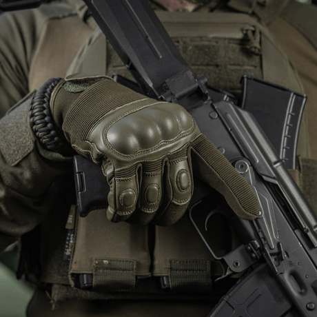 Person iført M-Tac Handsker Assault Tactical Mk.4 holder et våben; forstærkede knoepuder og velcro til sikker pasform