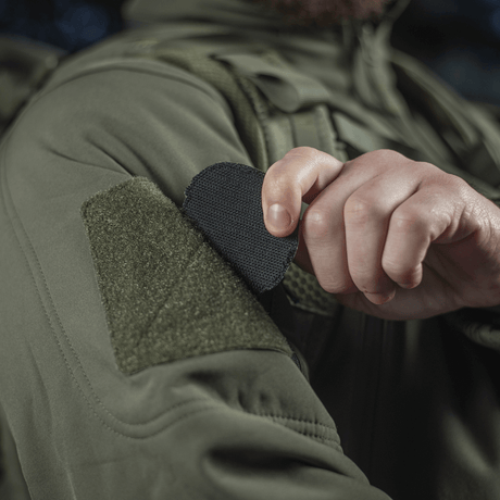 Henkilö kiinnittää M-Tac Patch Operator 3D PVC:n Velcro-kiinnityksellä takkiin, taktinen muotoilu, moraalimerkki, 6x7 cm, PVC.
