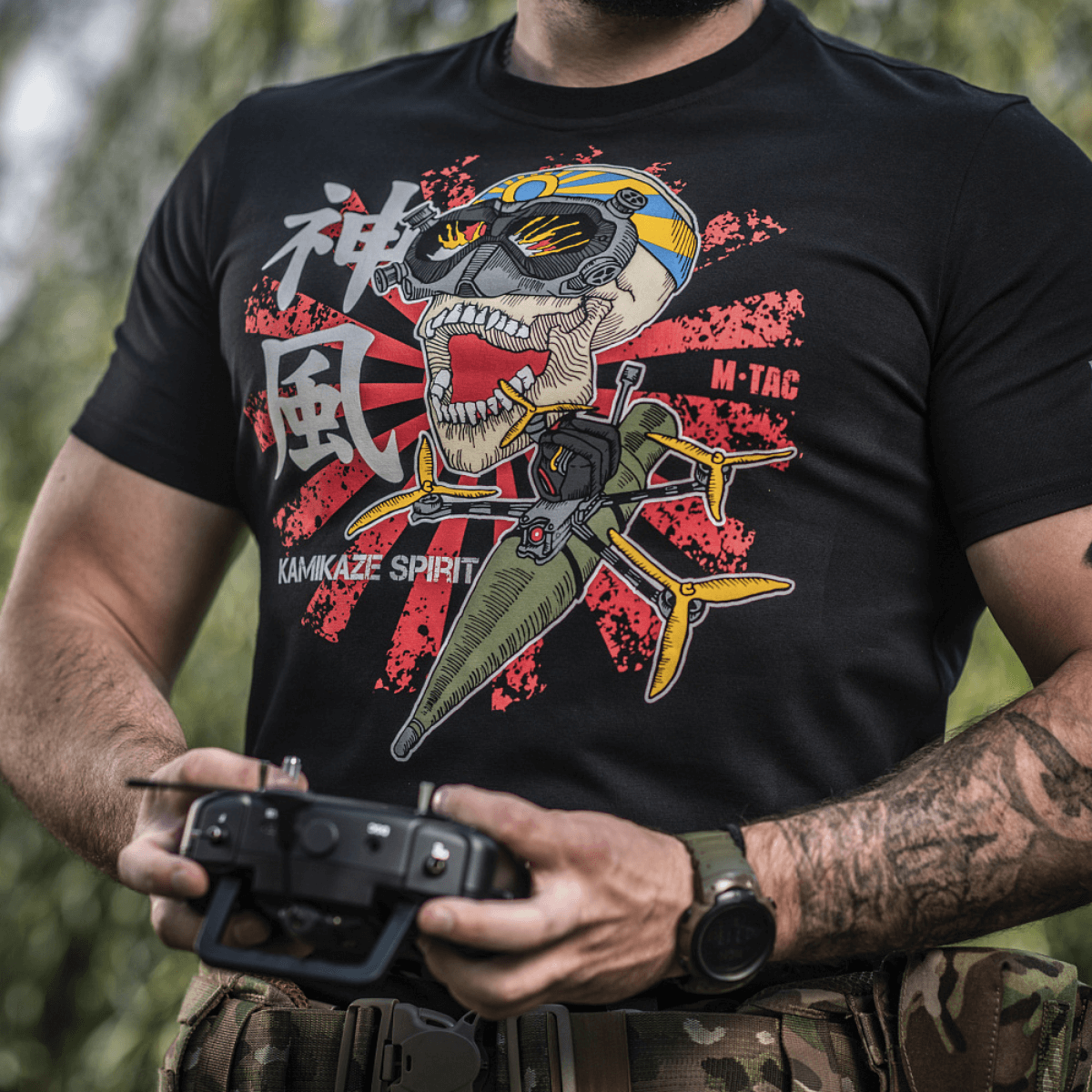 Mand iført M-Tac T-Shirt Kamikaze Spirit med FPV-design, holder en dronecontroller. Sort t-shirt med stiliseret kranieprint.