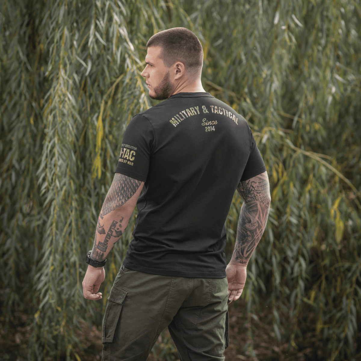 M-Tac T-Shirt Logo med militær camouflage og slogan "Born by Revolution – Hardened by War" i naturligt baggrundsmiljø.