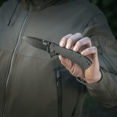 Henkilö pitää M-Tac Outdoor & Militær Type 7 -taittoveistä, musta terä ja ergonominen kahva, sopii taktiseen käyttöön.