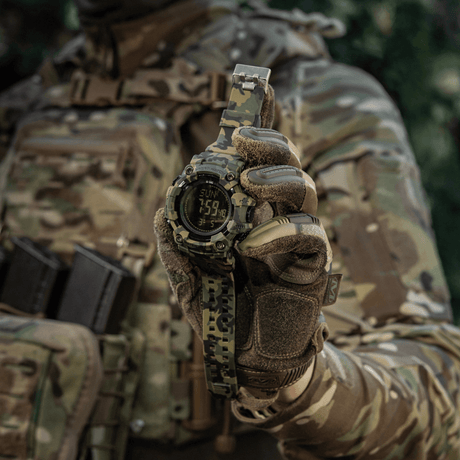 M-Tac Tactical Ur 2037 camo-kuosissa, pidetty taktisen varustuksen käyttäjän toimesta, näyttää kellonajan ja päivämäärän, sopii ulkoiluun ja arkeen.