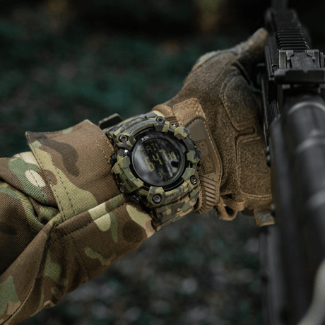 M-Tac Tactical Ur 2037 ranteessa maastovaatteiden kanssa, sopii ulko- ja taktiseen käyttöön, korostaa tukevaa muotoilua ja toiminnallisuutta.