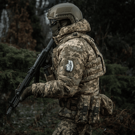 Sotilas maastopuvussa, M-Tac Patch M67 Kranaatti 3D PVC hihassa, aseistautuneena metsässä.
