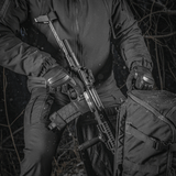 En person holder et våben ved siden af en M-Tac Rygsæk Gen.II Elite Large i en skovområde. Rygsækkens holdbare design er tydeligt.