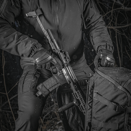 En person holder et våben ved siden af en M-Tac Rygsæk Gen.II Elite Large i en skovområde. Rygsækkens holdbare design er tydeligt.