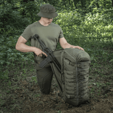 M-Tac Rygsæk Gen.II Elite Large i skoven med våben, viser holdbar konstruktion og funktionalitet til udendørs eventyr.