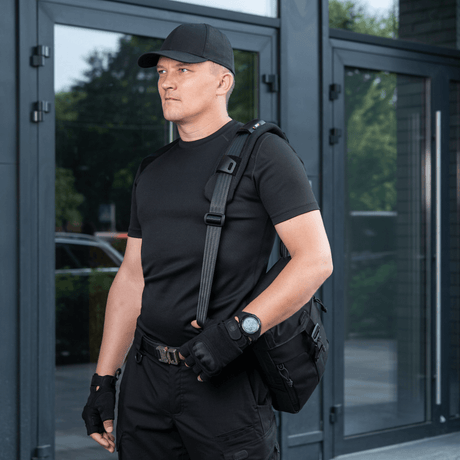 Mand bærer taske med M‑Tac Skulderremspude 50 mm Elite for øget komfort og støtte, iført sort tøj foran moderne bygning.