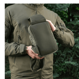 Person viser M-Tac Sling Pistoltaske Elite Hex udendørs i skovmiljø, iført jakke med lynlås.