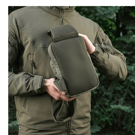 Person viser M-Tac Sling Pistoltaske Elite Hex udendørs i skovmiljø, iført jakke med lynlås.