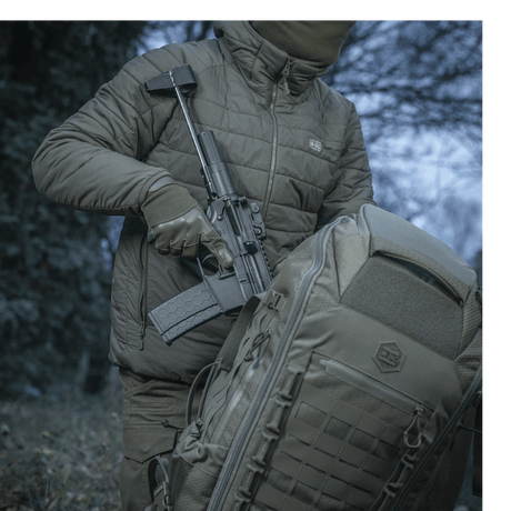 Person bærer våben ved siden af M-Tac Rygsæk Large Elite Hex Gen. IV i naturen, fremviser taskens holdbarhed og design.